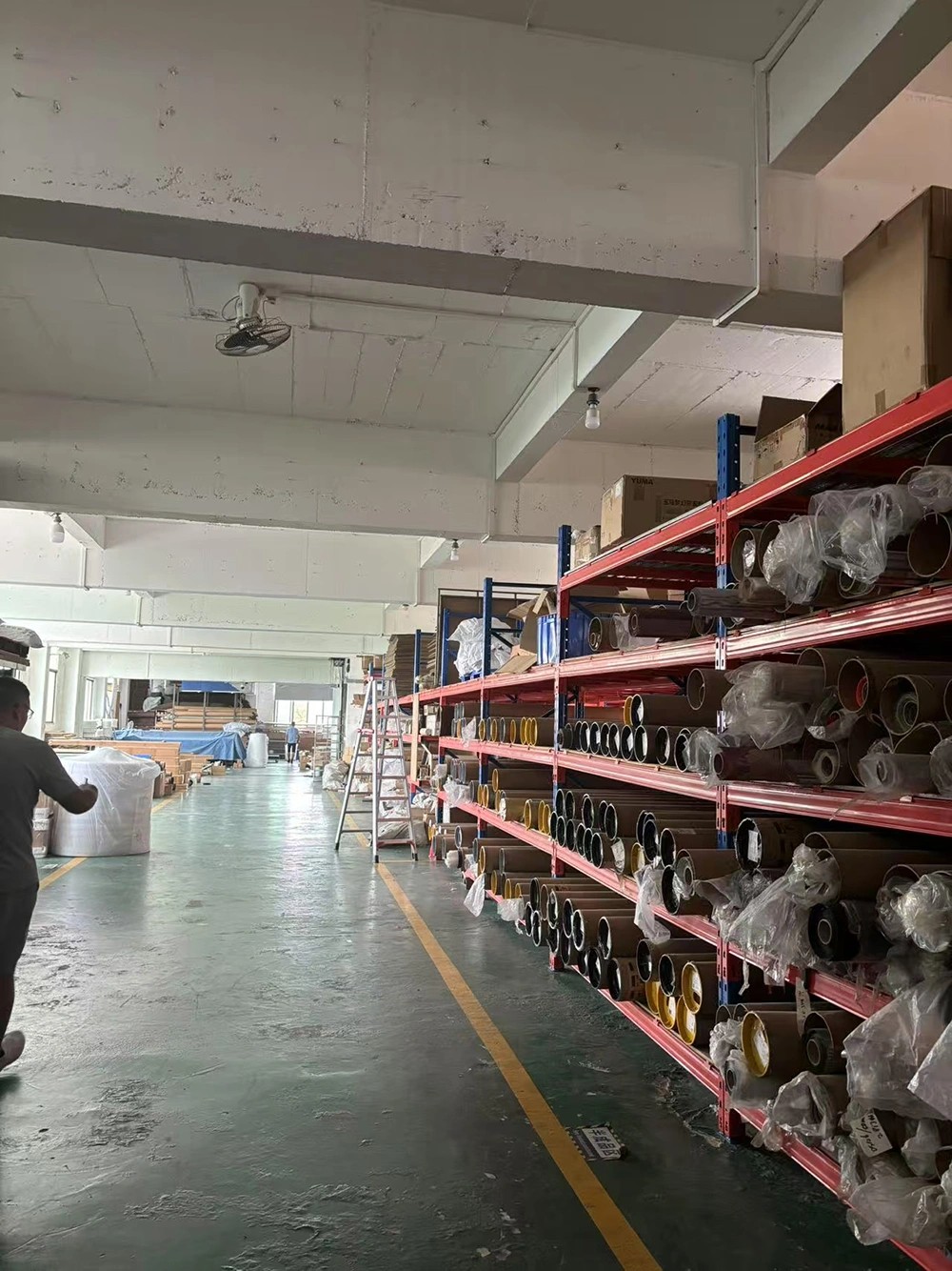 Organized factory aisle with stacked rolls of outdoor shade mesh on industrial racking.